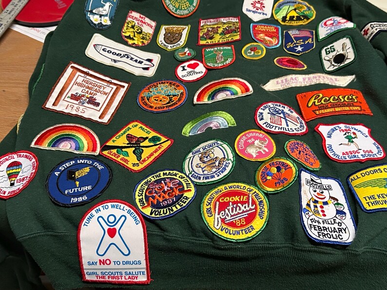 May include: A dark green sweatshirt covered in embroidered patches. Patches include rainbows, a Goodyear logo, and text that reads "Hershey Highmeadow Camp 1985". Other patches say "Cookie Festival" and "Say No To Drugs".