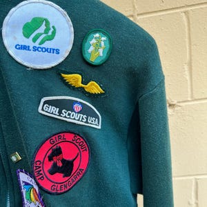 May include: Close-up of a dark green Girl Scout jacket adorned with various patches. Patches include the Girl Scouts logo, a floral design, and a red Camp Glengarra patch with a black dog illustration. The jacket has a zipper.