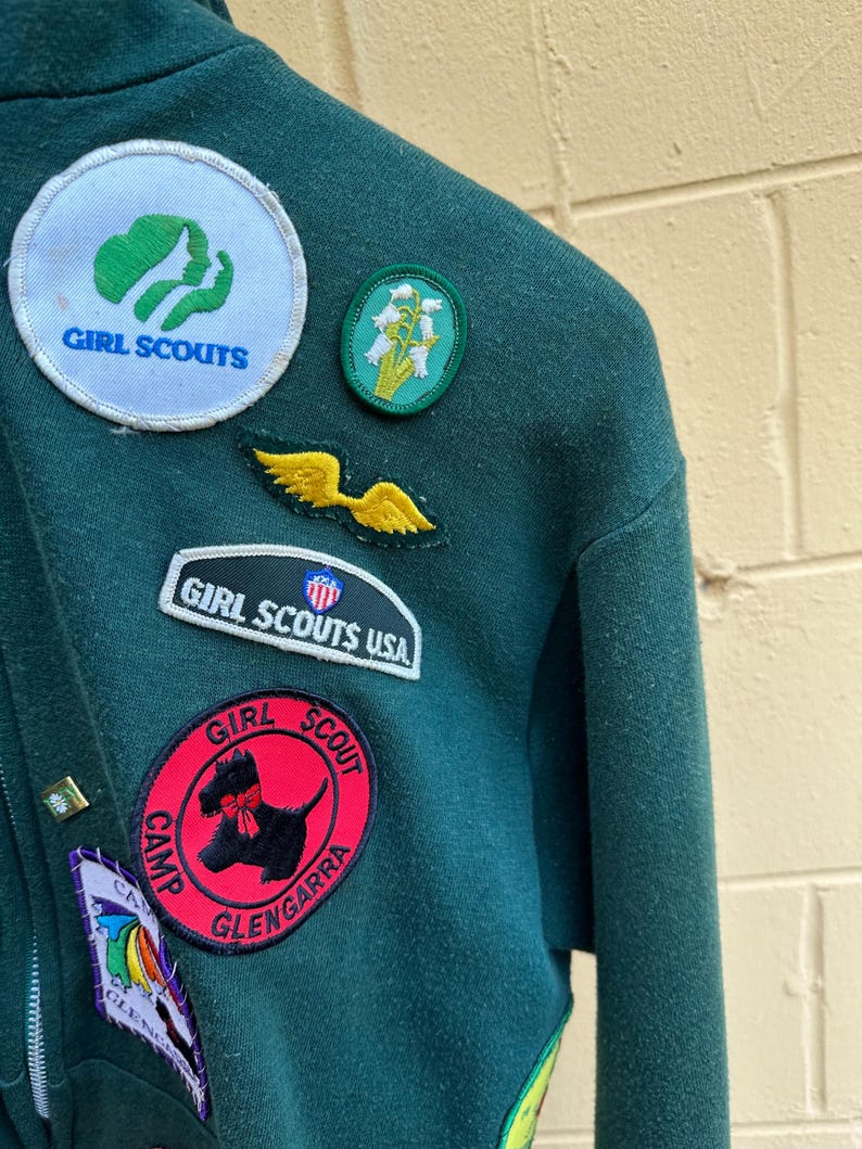 May include: Close-up of a dark green Girl Scout jacket adorned with various patches. Patches include the Girl Scouts logo, a floral design, and a red Camp Glengarra patch with a black dog illustration. The jacket has a zipper.