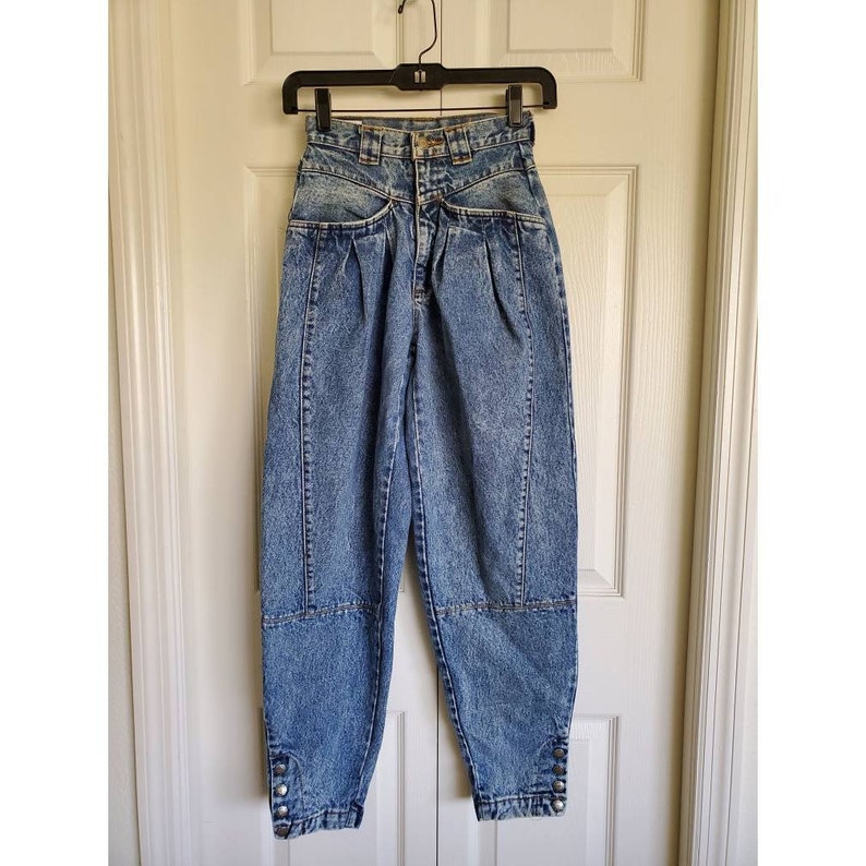 May include: High-waisted, light blue denim jeans with pleats and button details at the cuffs. The jeans have a relaxed fit and a cropped length.