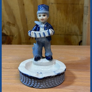 May include: A ceramic figurine of a boy playing an accordion. The boy is dressed in a blue uniform with a matching hat. The figurine is an ashtray with a white and blue decorative base.