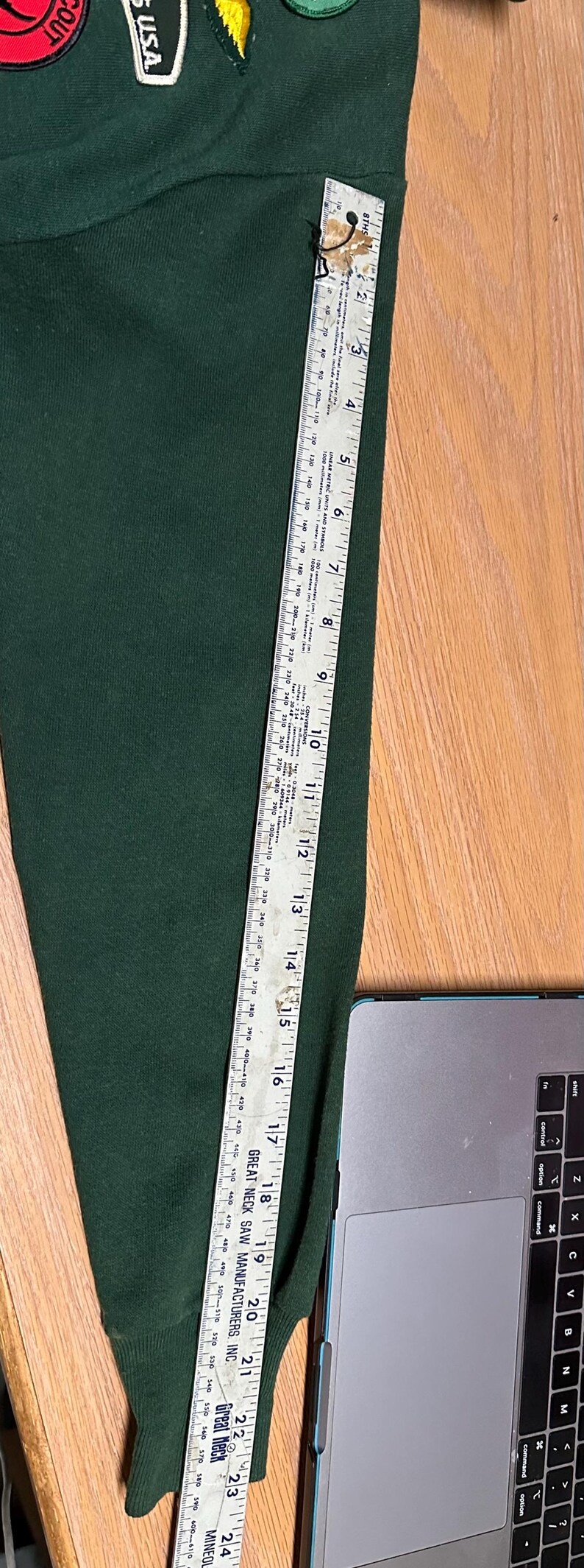 May include: Dark green sweater sleeve with patches. A ruler shows the sleeve is approximately 24 inches long. Patches include text that reads "USA" and "SCOUT".