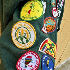 May include: Dark green sweatshirt covered in colorful patches. Patches include text like "Foothills Piper Event", "We Are Family", and "Franklin Pierce College 1987". Other patches feature a rainbow, a hot air balloon, and a butterfly.