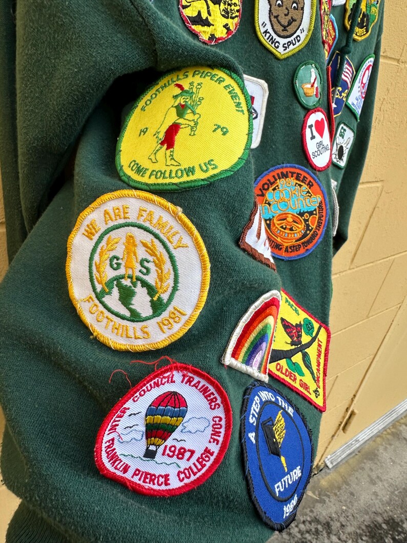 May include: Dark green sweatshirt covered in colorful patches. Patches include text like "Foothills Piper Event", "We Are Family", and "Franklin Pierce College 1987". Other patches feature a rainbow, a hot air balloon, and a butterfly.