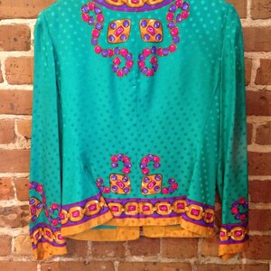 May include: A teal blue silk blouse with a gold chain and jewel pattern. The blouse has a polka dot pattern and a mandarin collar.