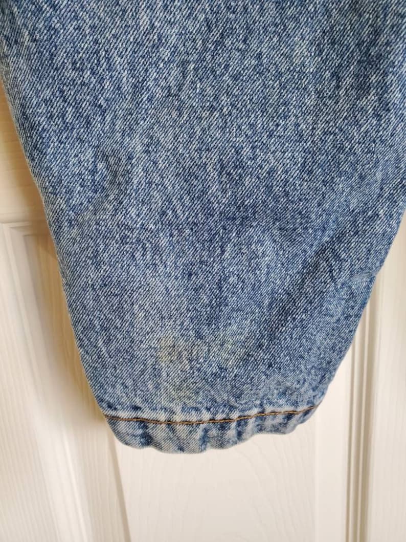 May include: Close-up of the bottom of a pair of light blue denim jeans. The denim has a faded, distressed look. The jeans have a brown seam around the hem.