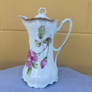 May include: White porcelain creamer with a gold trim and a floral design featuring pink roses and green leaves.