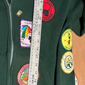 May include: Dark green zip-up jacket with multiple patches, including "Girl Scout" and "Scouts USA." Patches feature colorful designs and text. The jacket is approximately 25 inches long.