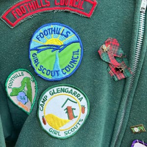 May include: Close-up of a dark green jacket with multiple patches and pins. Patches include "FOOTHILLS COUNCIL" and "FOOTHILLS GIRL SCOUT COUNCIL". Additional patches read "CAMP GLENGARRA GIRL SCOUTS" and "FOOTHILLS OF GS SINCE". A zipper runs down the right side.