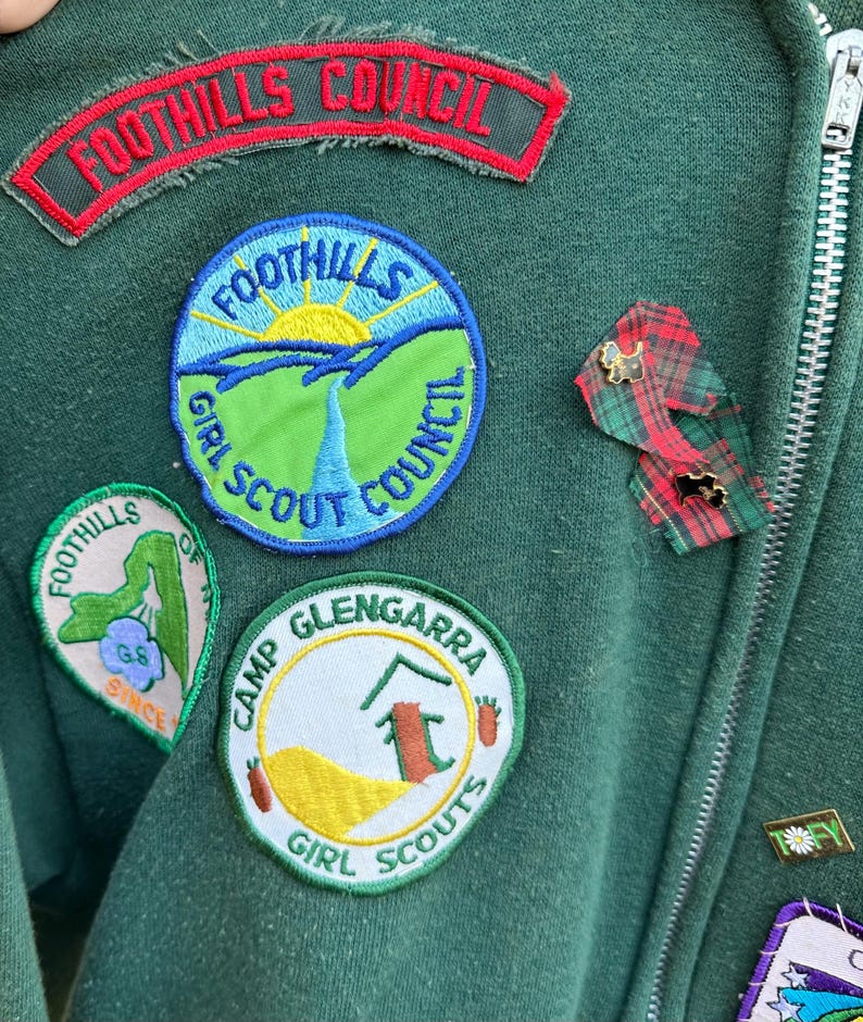 May include: Close-up of a dark green jacket with multiple patches and pins. Patches include "FOOTHILLS COUNCIL" and "FOOTHILLS GIRL SCOUT COUNCIL". Additional patches read "CAMP GLENGARRA GIRL SCOUTS" and "FOOTHILLS OF GS SINCE". A zipper runs down the right side.