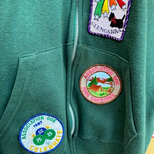 May include: A dark green zip-up hooded sweatshirt with multiple embroidered patches. Patches include a rainbow and dog design, a campsite scene, and a celebratory design with balloons. The sweatshirt has a front pocket and a silver zipper.