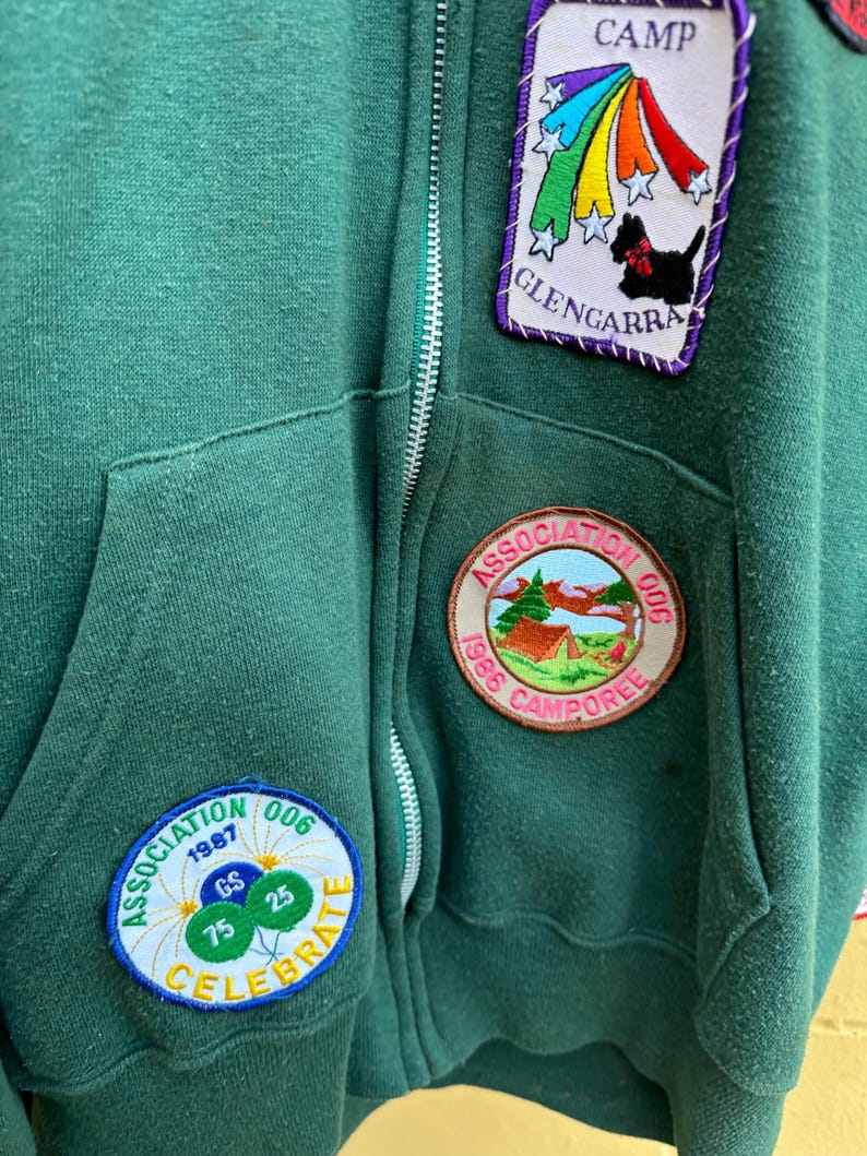 May include: A dark green zip-up hooded sweatshirt with multiple embroidered patches. Patches include a rainbow and dog design, a campsite scene, and a celebratory design with balloons. The sweatshirt has a front pocket and a silver zipper.
