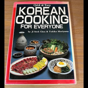 May include: A cookbook titled "Korean Cooking for Everyone" with a red border. The cover features a variety of Korean dishes, including a plate of sliced meat, vegetables, and a bowl of noodles with an egg. A vase and small bowls are also visible.