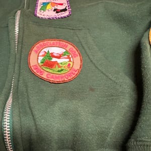 May include: Dark green zip-up hoodie with multiple patches. Patches include a Girl Scout Camp Glengarra badge with a black dog, a Camp Glengarra patch with a rainbow and dog, and a 1986 Camporee patch with a tent and trees.