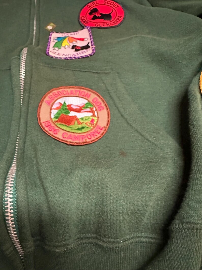May include: Dark green zip-up hoodie with multiple patches. Patches include a Girl Scout Camp Glengarra badge with a black dog, a Camp Glengarra patch with a rainbow and dog, and a 1986 Camporee patch with a tent and trees.