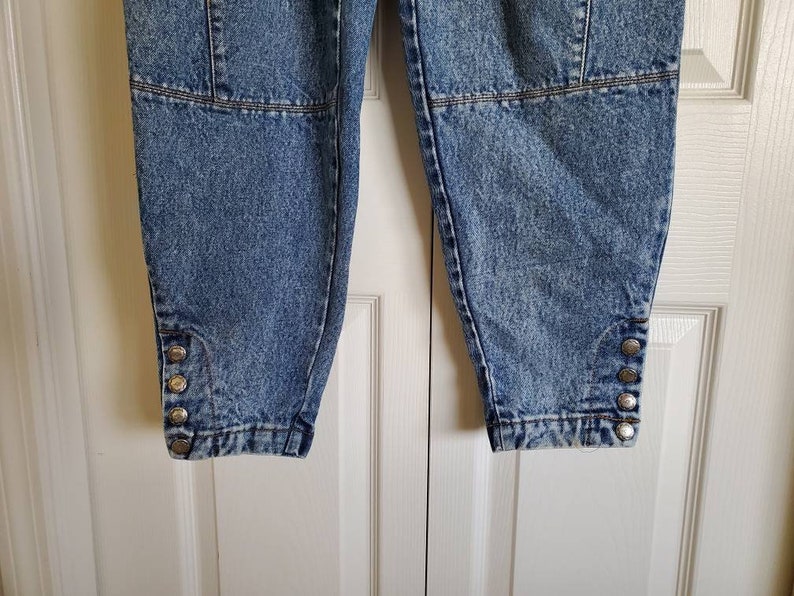 May include: A pair of light blue denim pants with a distressed look. The pants have a button detail on the bottom of each leg.