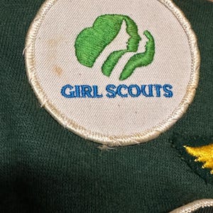 May include: A circular Girl Scouts patch with a green emblem and the words "GIRL SCOUTS" in blue. The patch is on a dark green fabric. The emblem features two profiles and a stylized leaf.