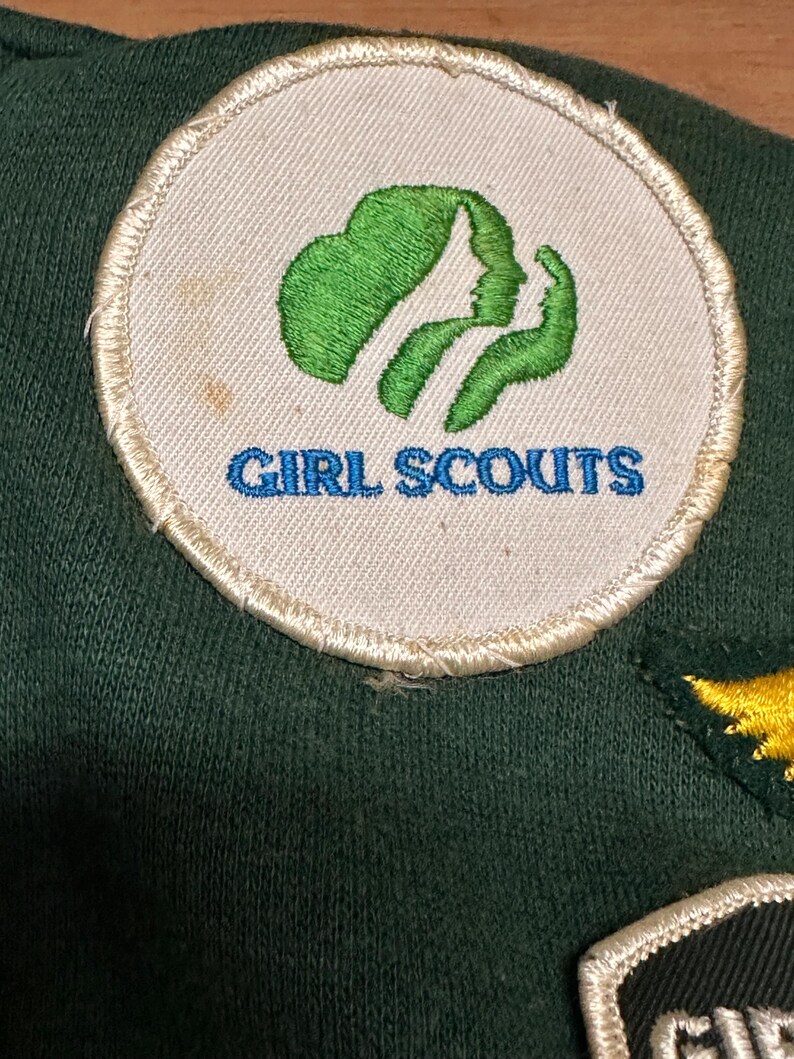 May include: A circular Girl Scouts patch with a green emblem and the words "GIRL SCOUTS" in blue. The patch is on a dark green fabric. The emblem features two profiles and a stylized leaf.