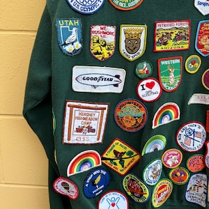 May include: Dark green sweatshirt adorned with a variety of colorful embroidered patches. Patches include references to the 1984 Olympics, Utah, Mt. Rushmore, Idaho, and the Girl Scouts. The sweatshirt features a vintage aesthetic with a mix of travel and activity-themed patches.