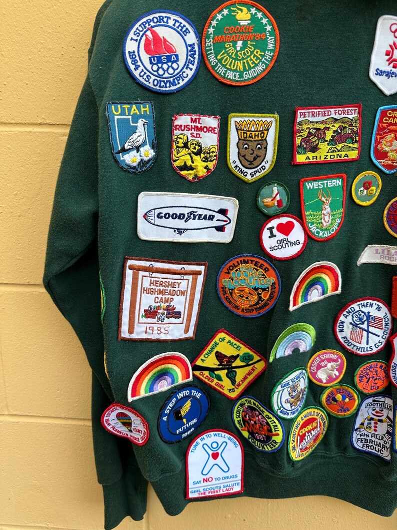 May include: Dark green sweatshirt adorned with a variety of colorful embroidered patches. Patches include references to the 1984 Olympics, Utah, Mt. Rushmore, Idaho, and the Girl Scouts. The sweatshirt features a vintage aesthetic with a mix of travel and activity-themed patches.