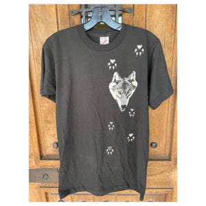 May include: Black t-shirt with a grey wolf graphic and paw prints. The wolf has yellow eyes.
