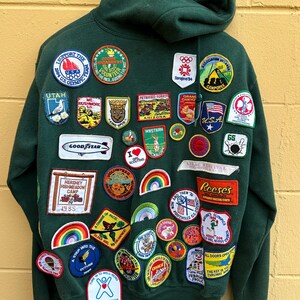 May include: Dark green hooded sweatshirt covered in colorful patches. Patches include the USA flag, Olympic logos, and various camp and event emblems. The sweatshirt has a hood and is made of a soft material.