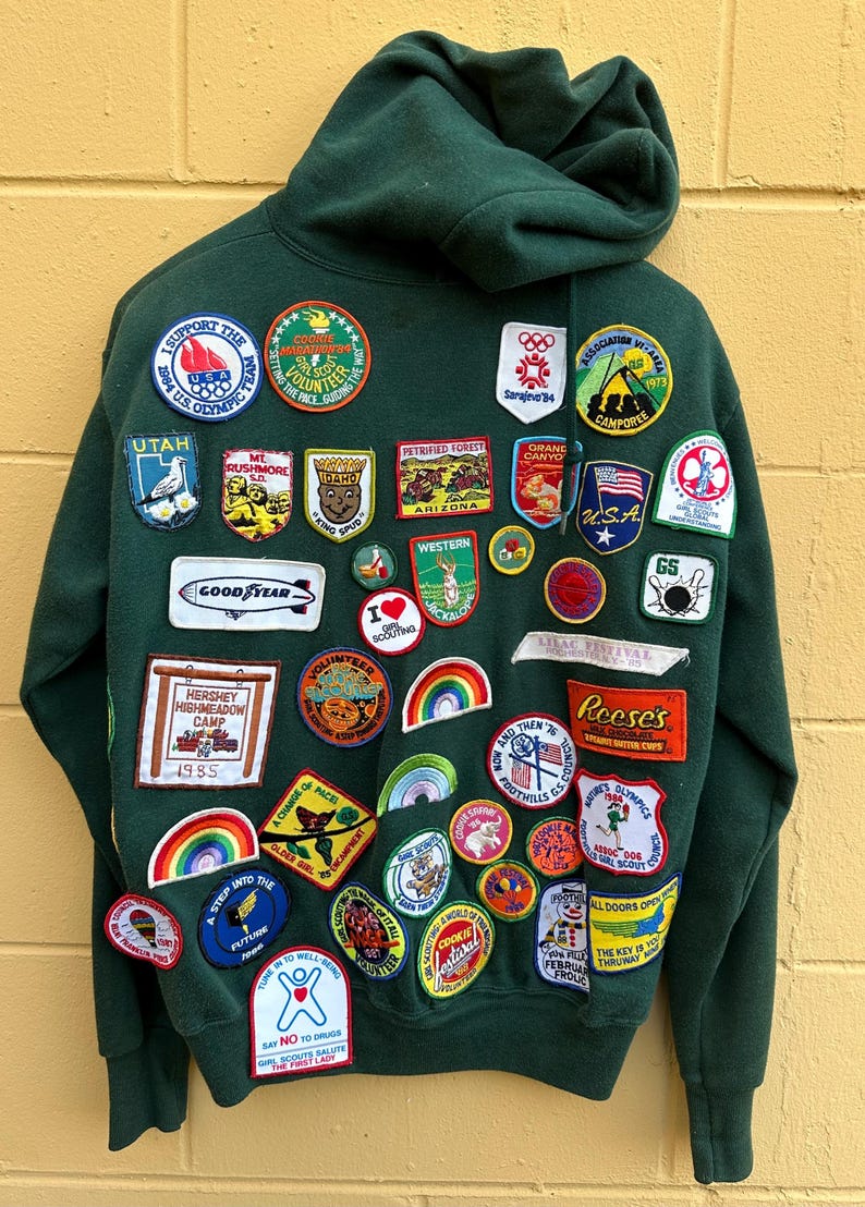 May include: Dark green hooded sweatshirt covered in colorful patches. Patches include the USA flag, Olympic logos, and various camp and event emblems. The sweatshirt has a hood and is made of a soft material.