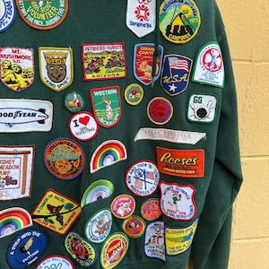 May include: A dark green hooded sweatshirt covered in colorful embroidered patches. The patches feature various designs, including rainbows, cartoon characters, and text such as "Cookie Marathon '84" and "Girl Scouts". The sweatshirt has a vintage aesthetic.