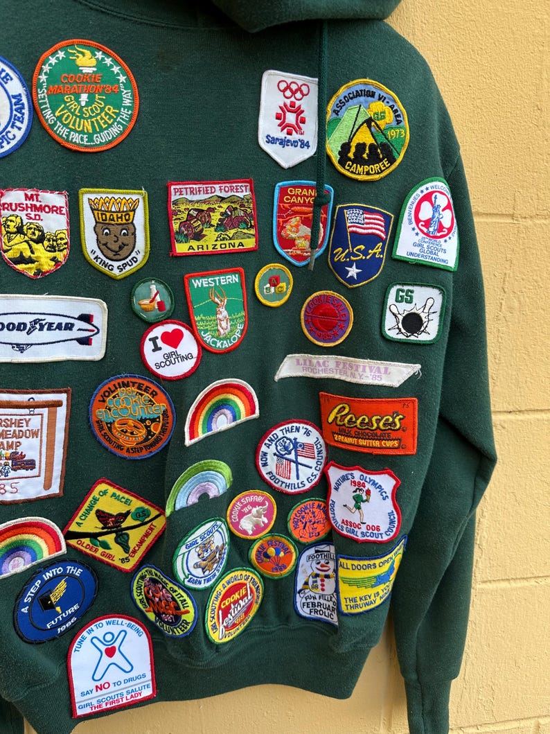 May include: A dark green hooded sweatshirt covered in colorful embroidered patches. The patches feature various designs, including rainbows, cartoon characters, and text such as "Cookie Marathon '84" and "Girl Scouts". The sweatshirt has a vintage aesthetic.