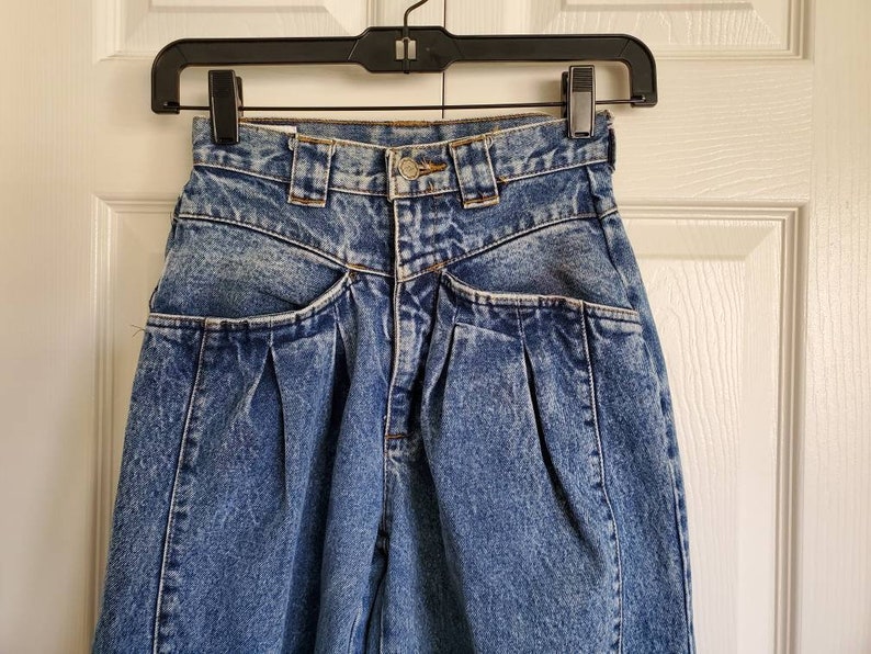 May include: A pair of blue denim jeans with pleats and multiple pockets. The jeans have a high waist and a relaxed fit.
