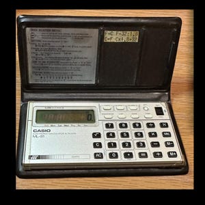 May include: A vintage Casio ML-81 electronic calculator and clock. The silver calculator has a digital display showing the time and date. It features a full numeric keypad and function buttons. The calculator is housed in a black case with a flip-up lid.