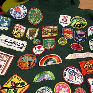 May include: A dark green hoodie covered in colorful embroidered patches. Patches include the U.S. Olympic Team, Girl Scout Marathon, Utah, Mt. Rushmore, Goodyear, Hershey Highmeadow Camp, and Reese's Peanut Butter Cups. The patches vary in shape and size.