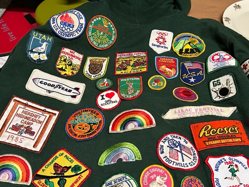 May include: A dark green hoodie covered in colorful embroidered patches. Patches include the U.S. Olympic Team, Girl Scout Marathon, Utah, Mt. Rushmore, Goodyear, Hershey Highmeadow Camp, and Reese's Peanut Butter Cups. The patches vary in shape and size.