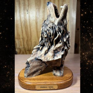 May include: A detailed sculpture of a howling wolf, carved from light brown wood. The wolf is mounted on a round, wooden base. The sculpture is labeled "PRIMAL SONG".