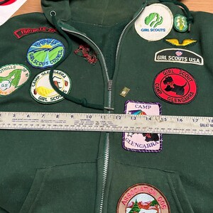 May include: Dark green hooded sweatshirt with a zipper closure and multiple patches. Patches include "Girl Scouts USA", "Camp Glengarra", and "Foothills Girl Scout Council". A ruler is placed across the front of the sweatshirt.