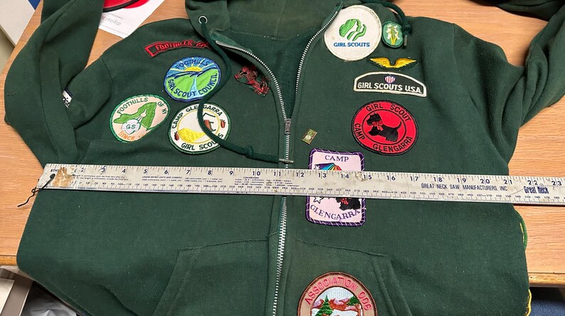May include: Dark green hooded sweatshirt with a zipper closure and multiple patches. Patches include "Girl Scouts USA", "Camp Glengarra", and "Foothills Girl Scout Council". A ruler is placed across the front of the sweatshirt.