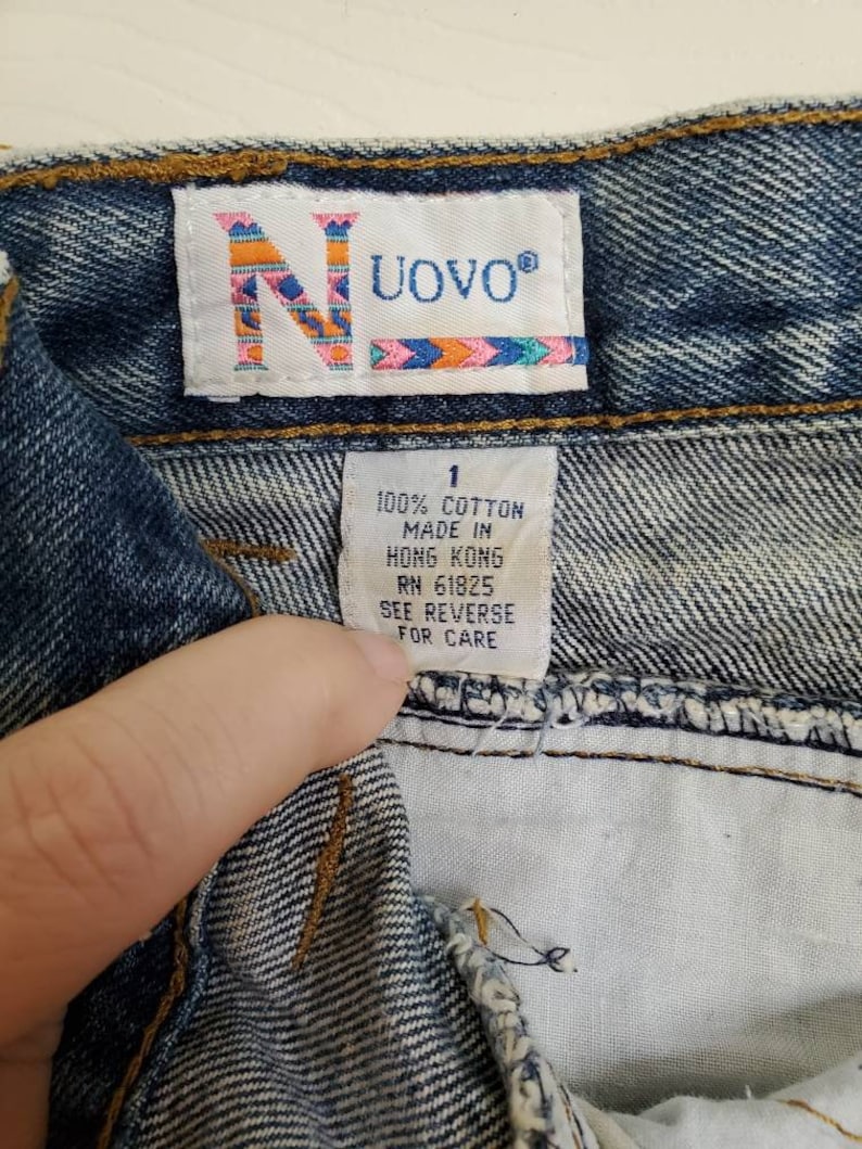 May include: A close-up of a blue denim jean waistband with a white tag that reads "UOVO&reg;" and "100% COTTON MADE IN HONG KONG RN 61825 SEE REVERSE FOR CARE".