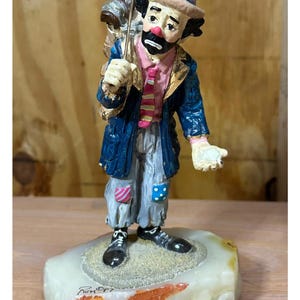 May include: A detailed figurine of a sad clown, dressed in a blue jacket, pink shirt, and gray pants with patches. The clown carries a bag and a golf club. The figurine stands on a stone base with a signature.
