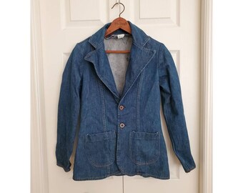 May include: A blue denim blazer with two front pockets and two buttons. The blazer is hanging on a wooden hanger against a white door.