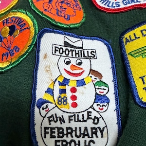 May include: A collection of embroidered patches with various designs. One patch features a snowman with a blue and yellow scarf, the word "FOOTHILLS" above, and the text "FUN FILLED FEBRUARY FROLIC" below. Other patches include text and cartoon graphics.