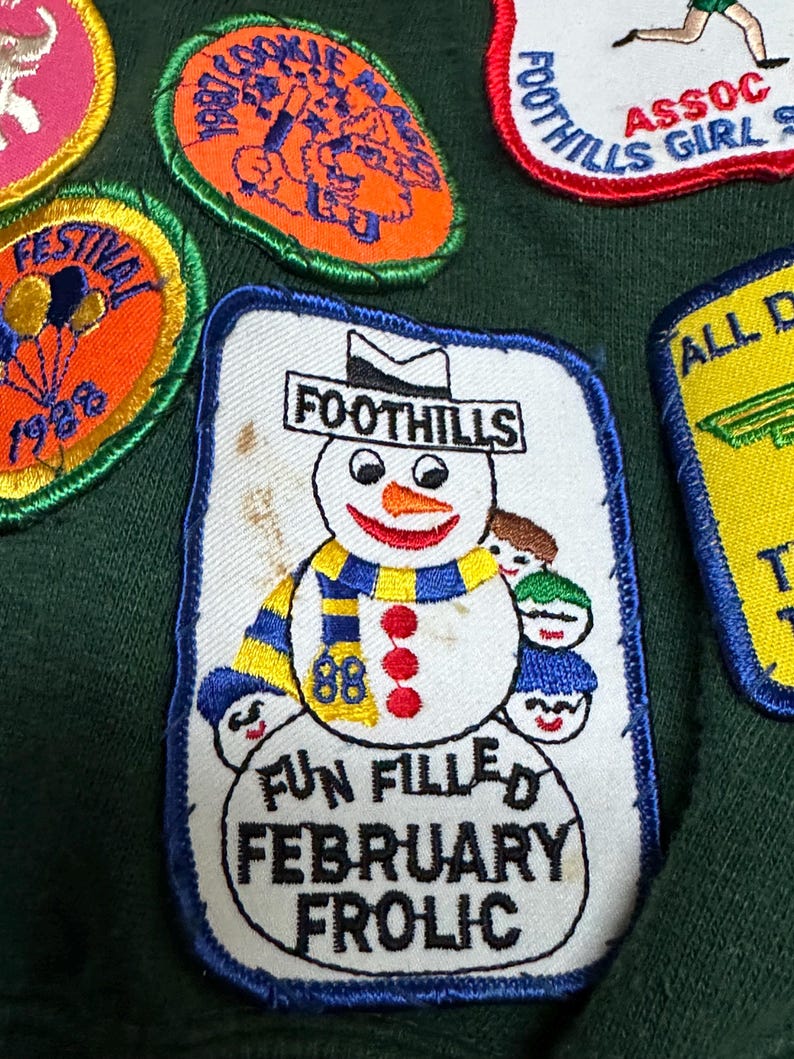 May include: A collection of embroidered patches with various designs. One patch features a snowman with a blue and yellow scarf, the word "FOOTHILLS" above, and the text "FUN FILLED FEBRUARY FROLIC" below. Other patches include text and cartoon graphics.