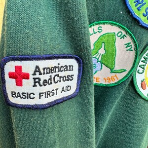 May include: Close-up of a dark green sweatshirt with several patches. The prominent patch features the words "American Red Cross BASIC FIRST AID" with a red cross symbol. Other patches include "GIRL SCOUTS" and a New York state outline.