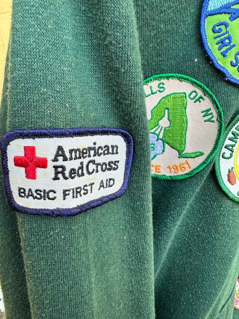 May include: Close-up of a dark green sweatshirt with several patches. The prominent patch features the words "American Red Cross BASIC FIRST AID" with a red cross symbol. Other patches include "GIRL SCOUTS" and a New York state outline.