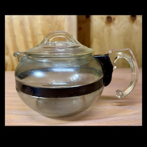May include: A vintage glass teapot with a clear glass lid and handle. The teapot has a silver-colored base and a dark brown band around the middle. The handle is curved and clear. The teapot is sitting on a wooden surface.