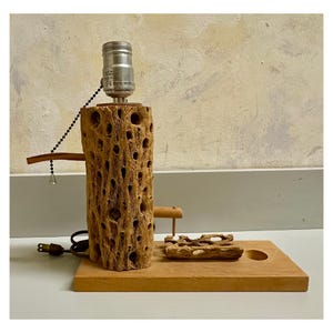 May include: A unique table lamp featuring a cylindrical, textured wooden base with numerous holes. The lamp includes a silver-colored bulb socket and a small wooden accent piece. The lamp sits on a rectangular wooden base with a small hole.