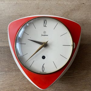 Op de afbeelding: Een vintage wandklok met een witte wijzerplaat, zwarte wijzers en een rood-wit driehoekig frame. De wijzerplaat heeft het woord "Junghans" en de cijfers 12 en 6. Retro design.