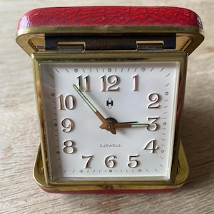 Op de afbeelding: Een vintage reiswekker met een rode kunstleren behuizing en een goudkleurig metalen frame. De witte wijzerplaat heeft zwarte cijfers, gouden wijzers en de tekst "2 JEWELS". De klok is open en toont de tijd.