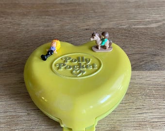 May include: A vintage Polly Pocket toy in a heart shape, colored in a pale yellow-green. The toy has the words "Polly Pocket" embossed on the top. Two small plastic figures are on top of the toy. One figure is a person lying down, and the other is a person riding a horse.