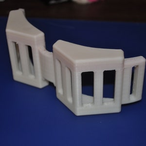 May include: White plastic 3D printed object with a curved shape and multiple rectangular openings. The object is designed with a repeating pattern of vertical bars.