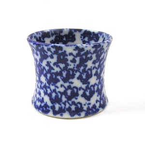 May include: A small, cylindrical ceramic cup featuring a white base with a dense, dark blue mottled or speckled pattern throughout the exterior surface. The vessel has a slightly concave, hourglass-shaped profile with a smooth, rounded rim.